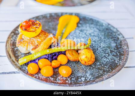 Gourmet-Fischgericht auf Teller mit gebratenen Kartoffeln, Spargel, Kirschtomaten, Purpurkohl und gegrillter Zitrone, präsentiert auf weißem Tisch. Stockfoto