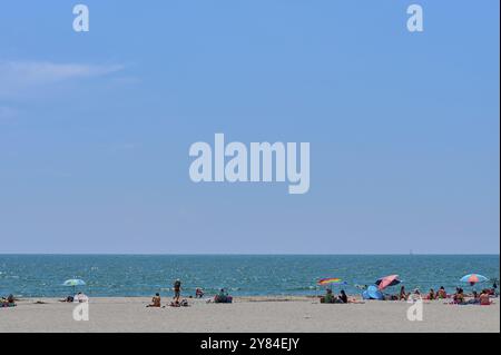 Strand mit Menschen und bunten Sonnenschirmen vor einem breiten, blauen Mittelmeer unter klarem Himmel, Saintes-Maries-de-la-Mer, Camargue, Frankreich, EUR Stockfoto