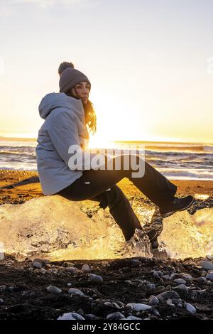 Porträt einer Frau, die bei Sonnenuntergang auf einem Stück Eis am Diamond Beach in Island sitzt Stockfoto