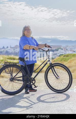 In einer bergigen Landschaft mit einem Dorf im Hintergrund fährt eine übergewichtige weißhaarige Frau ein Fahrrad im Retro-Stil Stockfoto