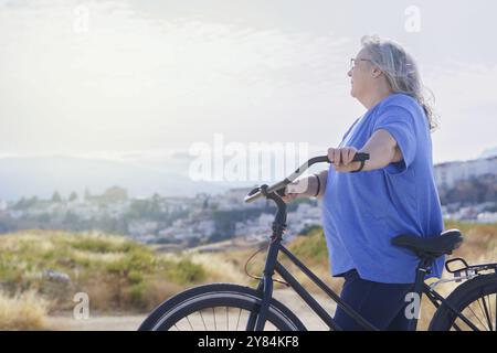 Eine übergewichtige reife Frau fährt ein schwarzes Fahrrad im Retro-Stil vor bewölkter Bergkulisse mit einem Dorf im Hintergrund Stockfoto