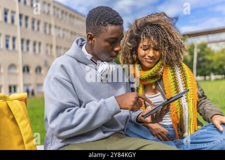 Lächelndes multiethnisches junges Paar, das ein Tablet benutzt, das außerhalb des Universitätsgeländes sitzt Stockfoto