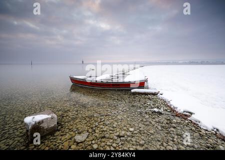 Ein schneebedecktes Rettungsboot liegt an einem bewölkten Wintertag am steinigen Ufer eines ruhigen Sees, Strandbad Hoernle, Konstanz, Bodensee, Baden-Wuertt Stockfoto