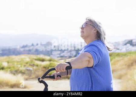 Übergewichtige weißhaarige Frau, die ein Fahrrad im Retro-Stil mit einer Berglandschaft mit einem Dorf im Hintergrund fährt Stockfoto