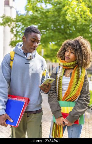 Vertikales Foto von zwei multiethnischen lateinischen und afrikanischen Studenten, die ihr Mobiltelefon nutzen, während sie auf dem Universitätsgelände spazieren Stockfoto