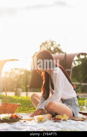 Schlankes, rothaariges Mädchen, das auf dem Boden im Park sitzt und den Sonnenuntergang betrachtet, während es ihr Picknick genießt. Stockfoto