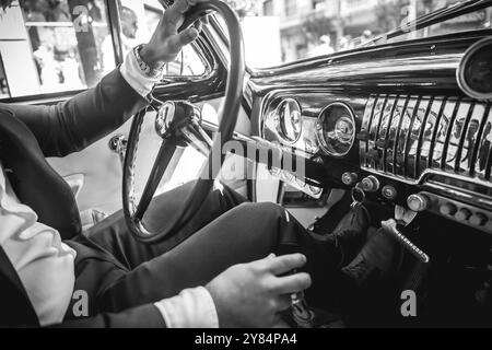 Ein Mann im Anzug fährt einen Oldtimer. Das Auto ist schwarz-weiß. Der Mann trägt eine Uhr Stockfoto