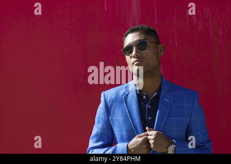 Vorderansicht eines jungen Mannes in einem blauen Anzug, der mit einer Sonnenbrille posiert und seine Jacke mit beiden Händen an einer roten Wand im Freien hält Stockfoto