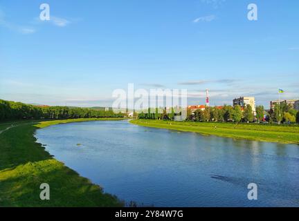 Malerischer Sommerblick auf die Stadt Uschhorod und den Fluss UZH, Transkarpaten, Ukraine. Ein ruhiger Fluss schlängelt sich durch üppige grüne Felder, die von Bäumen umgeben sind Stockfoto