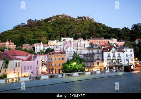 Maurisches Schloss Castelo dos Mouros Sintra Portugal // SINTRA, Portugal — die alte maurische Burg (Castelo dos Mouros) steht auf einem Hügel mit Blick auf die historische Stadt Sintra in der Abenddämmerung. Die Festung wurde im 8. Und 9. Jahrhundert von den Mauren erbaut, die die iberische Halbinsel besetzten. Sie besteht aus Steinmauern, die sich entlang des bergigen Geländes schlängeln. Die zum UNESCO-Weltkulturerbe gehörende Stadt Sintra breitet sich im Vordergrund mit ihrer unverwechselbaren Architektur und den roten Ziegeldächern aus, die in das sanfte Abendlicht getaucht sind. Sintra liegt etwa 25 Kilometer westlich von Lissabon und ist bekannt Stockfoto