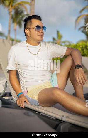 Entspannen auf einem Stuhl am Strand, junger lateinmann mit Sonnenbrille an einem sonnigen Tag, entspannter Lebensstil, Sommermode für Herren Stockfoto