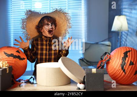 Süßer Junge, der wie Vogelscheuche gekleidet ist, mit festlichem Dekor und Geschenkboxen drinnen in der Nacht. Halloween-Feier Stockfoto