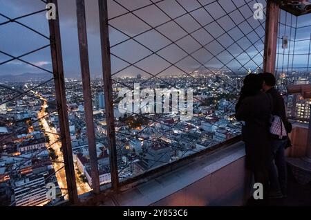 Beobachtungsdeck des Torre Latinoamericana Besucher Mexiko-Stadt Nacht | MEXIKO-STADT, Mexiko — Ein Besucher betrachtet die nächtliche Ausdehnung von Mexiko-Stadt durch den Drahtgitter-Sicherheitsbildschirm des Miradors, des höchsten öffentlichen Beobachtungsdecks im Centro Histórico in 166 m (545 ft) im 44. Stock des Torre Latinoamericana. Der Turm, der diesen Aussichtspunkt trägt, ist selbst das Wahrzeichen: auf 361 Betonpfählen gegründet, die bis zu einer Sandschicht 33 m (108 ft) unter dem weichen Seebett-Ton getrieben wurden, überstand er das katastrophale M8,1-Erdbeben von 1985 ohne erhebliche Schäden, während Hunderte von Gebäuden um ihn herum einstürzten. Stockfoto