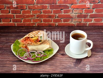 Ein Teller mit köstlicher saftiger Schawarma auf einem Salatblatt mit Zwiebeln und einer Tasse heißem, verpacktem schwarzem Tee auf einer Untertasse auf einem dunklen Holztisch. Nahaufnahme. Stockfoto