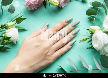 Eine wunderschön gepflegte Hand liegt auf einer türkisfarbenen Oberfläche, die mit langen, hellgrünen Nägeln verziert ist, umgeben von weißen Rosen und rosafarbenen Blüten, die schaffen Stockfoto