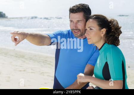 Ein Paar trägt Neoprenanzüge am Strand und zeigt nach vorne Stockfoto