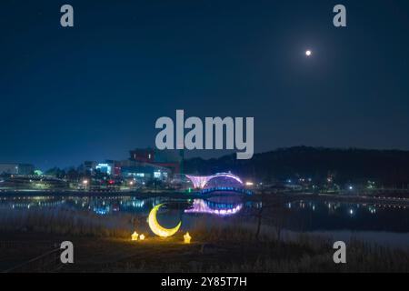 Ruhige Nacht im Gimpo Lake Park bei Vollmond. Die beleuchtete Brücke und die halbmondförmige Skulptur reflektieren auf ruhigem Wasser und schaffen eine friedliche Szene. Stockfoto