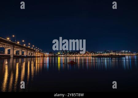 Dieses Foto zeigt einen Park bei Nacht, wo die Lichter der Stadt auf dem ruhigen Flusswasser reflektieren und die beleuchtete Brücke die ruhige Atmosphäre verstärkt. Stockfoto