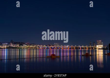 Dieses Foto zeigt einen nächtlichen Blick auf einen Park, in dem die Lichter der Stadt auf dem ruhigen Flusswasser reflektieren und die beleuchtete Brücke die ruhige Atmosphäre unterstreicht. Stockfoto
