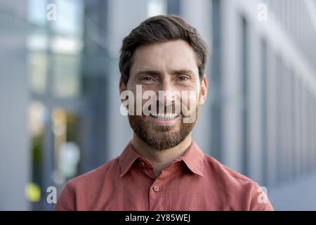 Ein Mann mit Bart und rotem Hemd lächelt. Er steht vor einem Gebäude Stockfoto