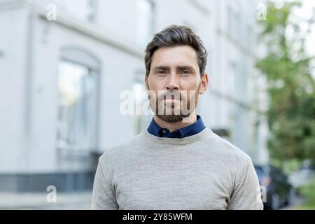 Ein Mann mit Bart und blauem Hemd steht vor einem Gebäude. Er trägt einen grauen Pullover Stockfoto