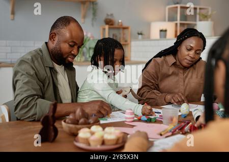 Afroamerikanische Familie genießt gemeinsame Zeit und kreiert Kunstprojekte zu Hause mit verschiedenen Vorräten und Snacks auf dem Tisch. Eltern und Tochter, die sich auf kreative Aktivitäten konzentrieren Stockfoto