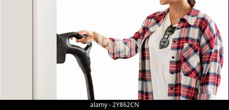 Isolierte weibliche Person in rotem kariertem Hemd mit Elektrostecker in der Hand an der Ladestation für Elektrofahrzeuge. Stockfoto