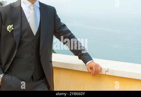 Der Bräutigam, der einen Anzug mit einem Boutonniere trägt, lehnt sich auf ein Geländer mit einem Ozean im Hintergrund Stockfoto