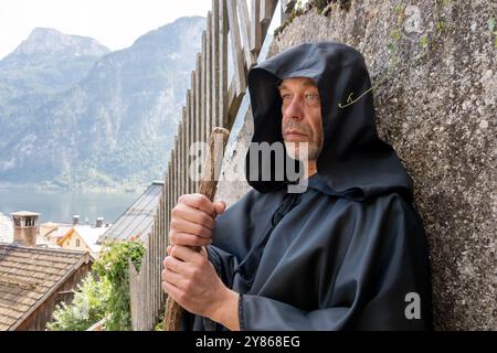 Ein älterer Mönch in einem schwarzen Gewand mit einem Personal steht an der Wand. Stockfoto