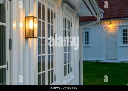 Nahaufnahme Details der weißen, holzgetäfelten Sommerhausfassade am Abend mit warmem Licht von einer Außenwandleuchte mit einem anderen Haus im Hintergrund. Stockfoto