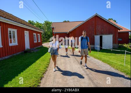 Eine Gruppe junger Erwachsener und ein Seniorin spazieren auf einer Schotterstraße zwischen roten, hölzernen Bauernhäusern auf der Insel Jomfruland an einem sonnigen Tag in Telemark, Norwegen. Stockfoto
