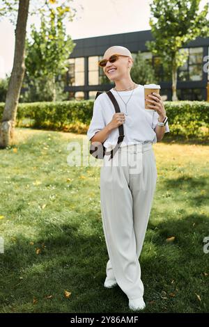 Eine kahlköpfige Frau lächelt hell, während sie mit einem Drink in der Hand durch einen lebhaften Park schlendert. Stockfoto