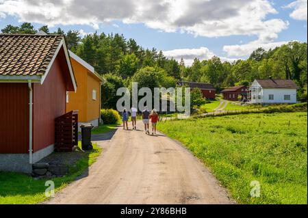 Urlauber spazieren an einem Bauernhof auf einer gewundenen Gasse mit Weide, idyllischen traditionellen Holzhäusern und Wäldern auf der Insel Skåtøy, Telemark, Norwegen. Stockfoto
