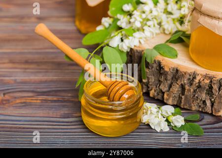 Süßes Honigglas umgab die Akazienblüten des Frühlings. Honig fließt aus einem Löffel in ein Glas. Gläser von klarem frischen Akazienhonig auf Holzgrund. Stockfoto