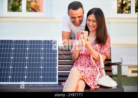 Lächelnde Frau, die ihrem Mann zeigt, wie man Solarladegerät benutzt. Junger Mann begeistert von erneuerbaren Energiequellen. Niedliches Paar, das auf das Telefon schaut und neben einem tragbaren Solarpanel sitzt. Stockfoto