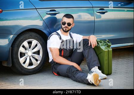 Tankstellenarbeiter macht nach einem geschäftigen Morgen eine Pause. Müder, aber zuversichtlicher Fahrer wartet auf den nächsten Kunden. Männlicher Arbeiter in Sonnenbrille und Overall, der auf dem Boden sitzt und sich auf Auto und Kanister stützt. Stockfoto