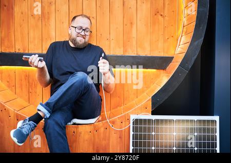 Bärtiger Mann, der Smartphone mit Photovoltaik-Sonnenkollektor verbindet. Integration nachhaltiger erneuerbarer Energien in den Alltag, demonstrieren die praktische Nutzung von Solarstrom für Ladegeräte. Stockfoto