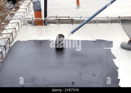 Arbeiter, der auf einer Baustelle eine bituminöse Grundierung auf eine Betonplatte aufträgt, bevor er bituminöse Abdichtungen verlegt Stockfoto