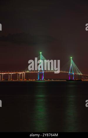 Ein nächtlicher Blick auf eine wunderschön beleuchtete Brücke mit blauen und violetten Lichtern, die auf dem ruhigen Wasser reflektieren und eine ruhige Atmosphäre schaffen. Stockfoto