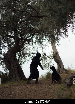 Zwei Figuren in Halloween-Kostümen stehen in einem Wald, eine hält eine Sense. Die dunkle Kleidung und die Umgebung zwischen alten Bäumen geben der Szene eine gruselige Atmosphäre Stockfoto