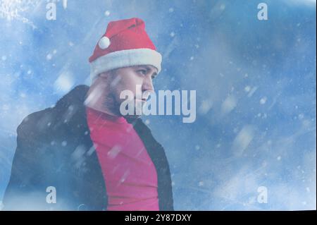 Ein bärtiger Mann mit Weihnachtsmann-Hut und rotem Hemd steht vor einem verschneiten, winterlichen Hintergrund. Die sanften Blautöne und der Schneefall deuten auf eine Weihnachtszeit hin Stockfoto