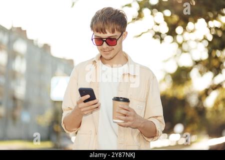 Lächelnder junger Mann, der auf dem Smartphone schreibt und Kaffee in der Morning City Street hält. Mit Beigefarbenem Hemd, Sonnenbrille Stockfoto