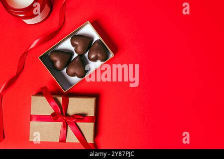 Eine Geschenkbox mit herzförmigen Pralinen, verziert mit einem roten Band auf einem leuchtend roten Hintergrund mit einer Kerze Stockfoto