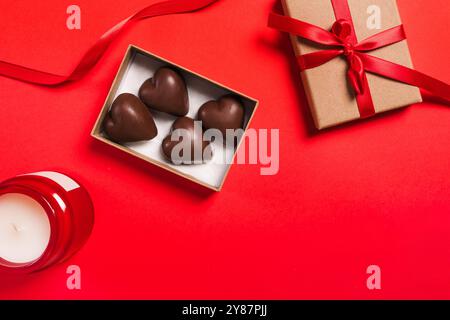 Eine Geschenkbox mit herzförmigen Pralinen, verziert mit einem roten Band auf einem leuchtend roten Hintergrund mit einer Kerze Stockfoto