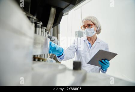 Schuss aus der Pillenpresse einer weiblichen Prozesstechnikerin in steriler Klassenbekleidung, die ein digitales Tablet hält, während sie Produktionsanlagen in der Pharmafabrik, Kopierraum, inspiziert Stockfoto