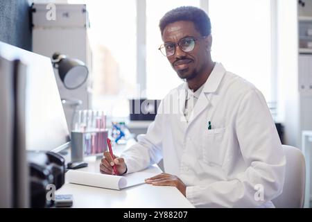 Mittleres Porträt von afroamerikanischem männlichen Medizinwissenschaftler, der in die Kamera lächelt, während er Forschungsaufzeichnungen im Notizblock macht, sitzend am Arbeitstisch im Labor, Kopierraum Stockfoto