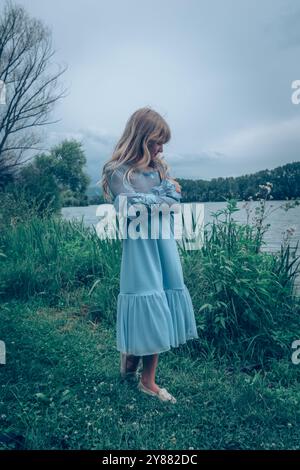 Schönes Mädchen in blauem Kleid auf grüner Wiese am Fluss auf dem Land Stockfoto