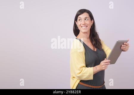 Eine lächelnde Frau in lässiger Kleidung hält eine Tafel und blickt auf ihre Seite. Sie trägt einen gelben Pullover über einem schwarzen Hemd und steht gegen eine neutrale Ba Stockfoto