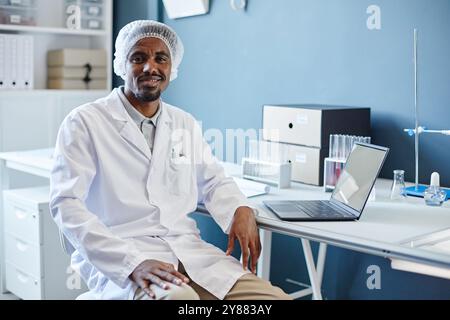 Mittleres Porträt Aufnahme des afroamerikanischen männlichen Laborchemikers in medizinischem Haarnetz lächelnd in die Kamera, während er am Arbeitstisch im Labor des wissenschaftlichen Forschungsdienstes sitzt, Kopierraum Stockfoto