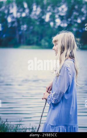 Wunderschönes bezauberndes göttliches Mädchen in blauem Kleid am Teich auf dem Land, magische Landschaft Stockfoto